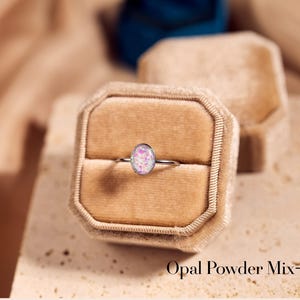 May include: A silver ring with an oval, pink and white opal stone, set in a velvet ring box. The text "Opal Powder Mix-In" is visible at the bottom of the image. The ring is a piece of jewelry.