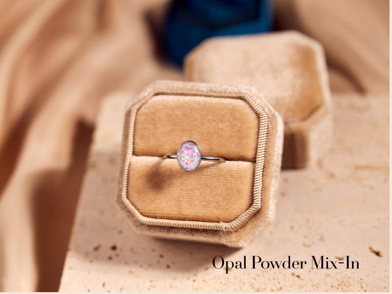 May include: A silver ring with an oval, pink and white opal stone, set in a velvet ring box. The text "Opal Powder Mix-In" is visible at the bottom of the image. The ring is a piece of jewelry.