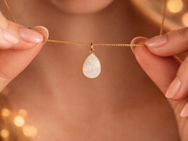 May include: A gold-colored necklace with a teardrop-shaped pendant. The pendant is white with a pearlescent sheen. The necklace is held by a person's hands, showcasing the delicate chain and pendant design. The jewelry is a necklace.