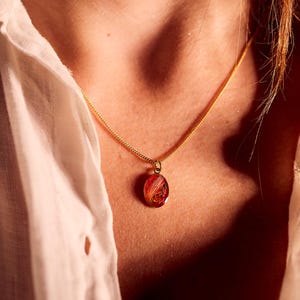 May include: A gold-colored chain necklace with a small, oval pendant. The pendant is a translucent red with embedded gold and orange accents. The necklace is worn against a white shirt.