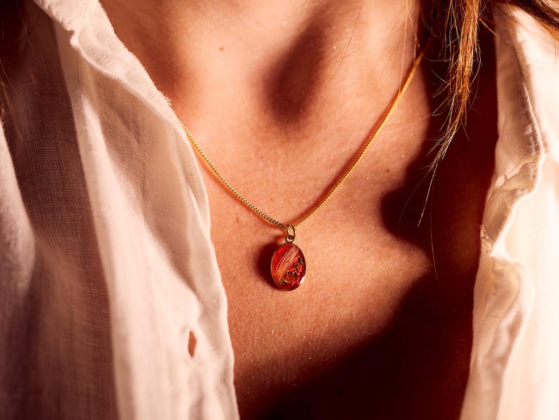 May include: A gold-colored chain necklace with a small, oval pendant. The pendant is a translucent red with embedded gold and orange accents. The necklace is worn against a white shirt.