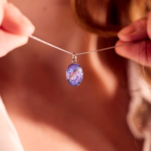 May include: A silver necklace with an oval pendant. The pendant features a vibrant mix of purple, pink, and white colors, creating a galaxy-like effect. The necklace is being held up, showcasing the pendant's design.
