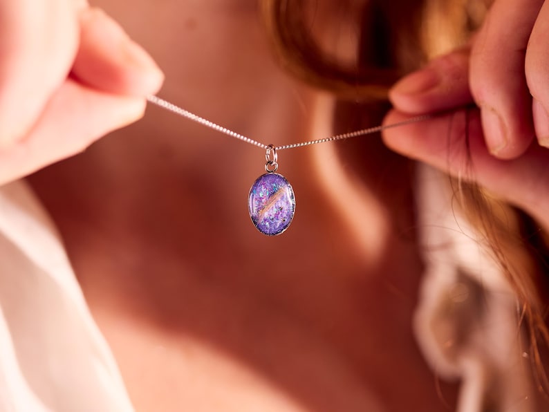 May include: A silver necklace with an oval pendant. The pendant features a vibrant mix of purple, pink, and white colors, creating a galaxy-like effect. The necklace is being held up, showcasing the pendant's design.