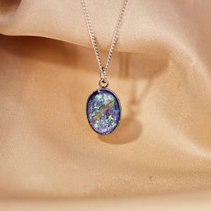 May include: A silver necklace with an oval pendant. The pendant features a blue and green design with a gold-colored accent. The necklace is displayed on a beige fabric background.