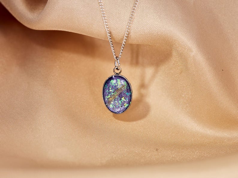 May include: A silver necklace with an oval pendant. The pendant features a blue and green design with a gold-colored accent. The necklace is displayed on a beige fabric background.