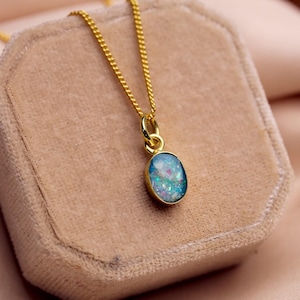 May include: A gold-colored necklace with a small, oval pendant. The pendant has a blue and white iridescent stone set in a gold bezel. The necklace is displayed on a beige velvet jewelry box.