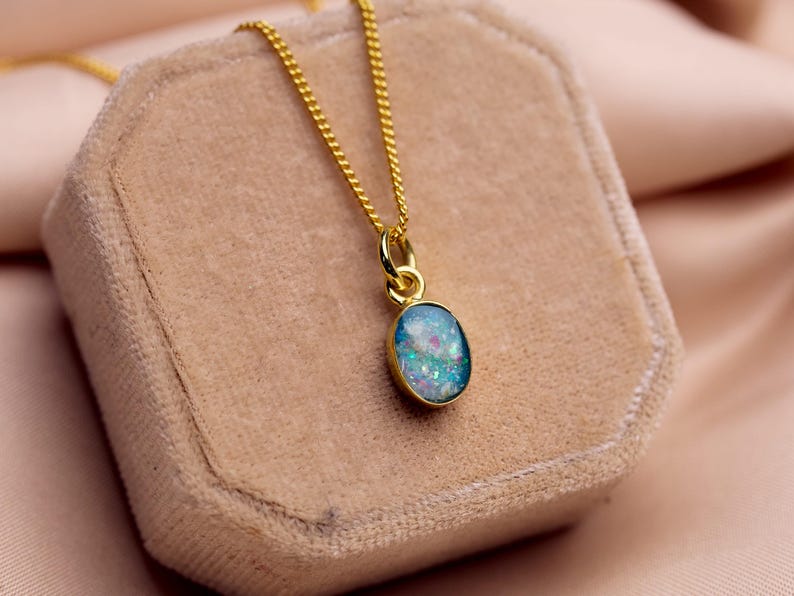May include: A gold-colored necklace with a small, oval pendant. The pendant has a blue and white iridescent stone set in a gold bezel. The necklace is displayed on a beige velvet jewelry box.