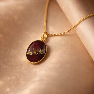 May include: A gold-colored necklace with an oval pendant. The pendant features a deep red background with gold leaf accents. The necklace is displayed on a soft, beige fabric.