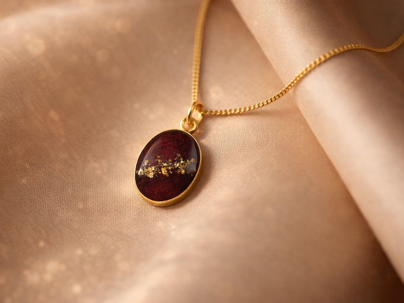 May include: A gold-colored necklace with an oval pendant. The pendant features a deep red background with gold leaf accents. The necklace is displayed on a soft, beige fabric.