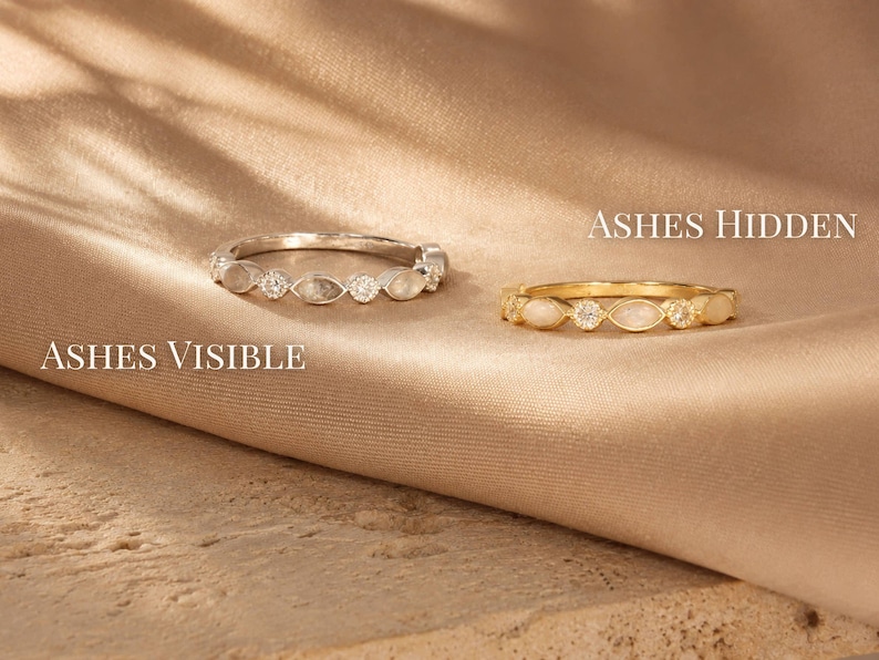 May include: Two rings, one silver and one gold, are displayed on a tan fabric. The silver ring, labeled "Ashes Visible," features alternating clear and opaque stones. The gold ring, labeled "Ashes Hidden," has a similar design. The text "Ashes Hidden" and "Ashes Visible" are also visible.