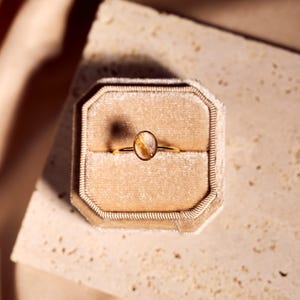 May include: A gold ring with an oval-shaped, amber-colored stone, set in a velvet ring box. The ring is centered in the frame, with the box resting on a textured surface. The background features soft, neutral tones.