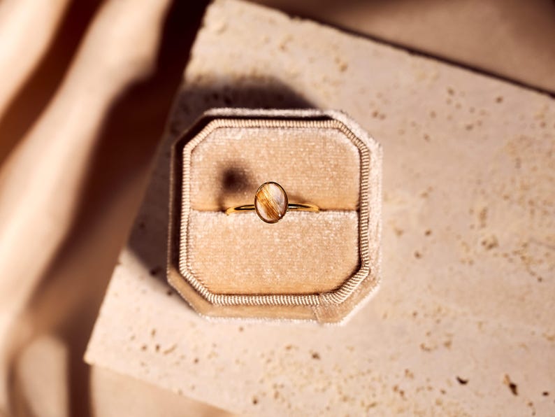 May include: A gold ring with an oval-shaped, amber-colored stone, set in a velvet ring box. The ring is centered in the frame, with the box resting on a textured surface. The background features soft, neutral tones.