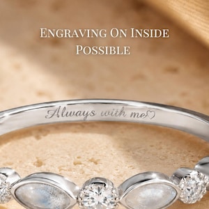 May include: Close-up of a silver ring with a delicate design. The ring features alternating clear and opalescent stones. Engraved on the inside is the phrase "Always with me" with a heart symbol. The text "Engraving on inside possible" is visible.