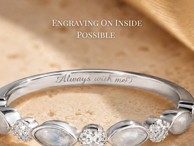 May include: Close-up of a silver ring with a delicate design. The ring features alternating clear and opalescent stones. Engraved on the inside is the phrase "Always with me" with a heart symbol. The text "Engraving on inside possible" is visible.