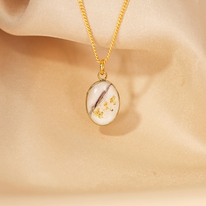 May include: A gold-colored necklace with a pendant featuring a white oval design with a dark brown streak and gold flecks. The necklace hangs against a beige fabric background.