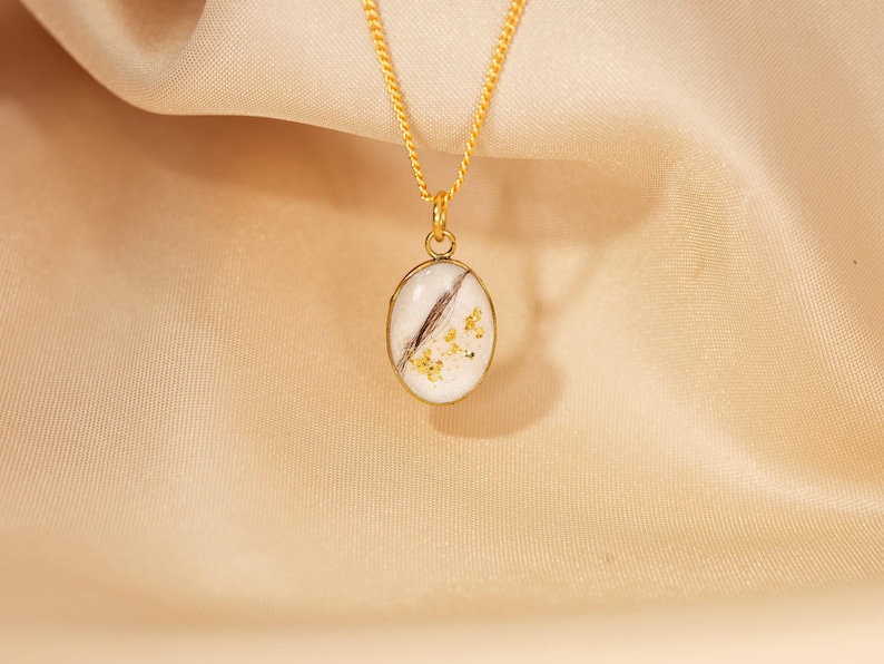 May include: A gold-colored necklace with a pendant featuring a white oval design with a dark brown streak and gold flecks. The necklace hangs against a beige fabric background.