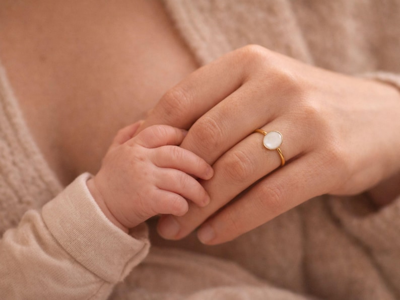 May include: A gold ring with a white oval stone is worn on a hand, held by a baby's hand. The ring is the focal point, set against a soft, neutral background. The baby's sleeve is a matching neutral color.