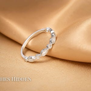 May include: A silver ring with alternating oval-shaped white stones and small clear stones. The ring is set against a gold satin fabric. The text "ASHES HIDDEN" is at the bottom of the image.