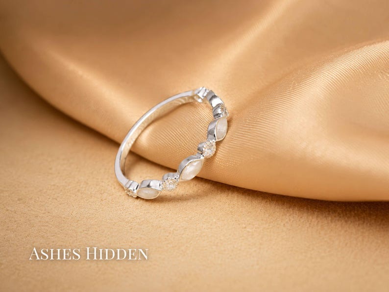May include: A silver ring with alternating oval-shaped white stones and small clear stones. The ring is set against a gold satin fabric. The text "ASHES HIDDEN" is at the bottom of the image.