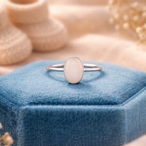May include: A silver ring with an oval, off-white stone is displayed on a blue velvet box. The ring is the focal point, with soft lighting and a blurred background that includes baby booties and floral accents. The ring's design is simple and elegant.