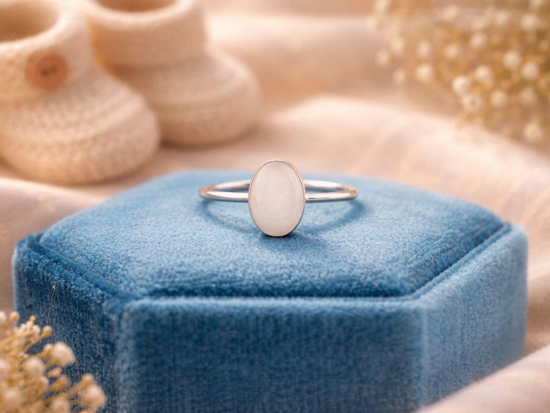 May include: A silver ring with an oval, off-white stone is displayed on a blue velvet box. The ring is the focal point, with soft lighting and a blurred background that includes baby booties and floral accents. The ring's design is simple and elegant.