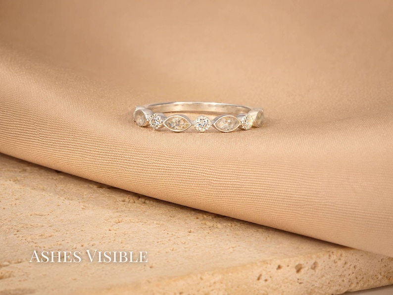 May include: A silver ring with alternating marquise-shaped and round clear stones. The ring is set against a tan fabric and a textured beige surface. The text "Ashes Visible" is at the bottom.