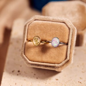May include: Two oval-shaped rings displayed in a velvet ring box. One ring has a gold band with a gold-colored stone, and the other has a silver band with a light purple and pink stone. The box is beige.
