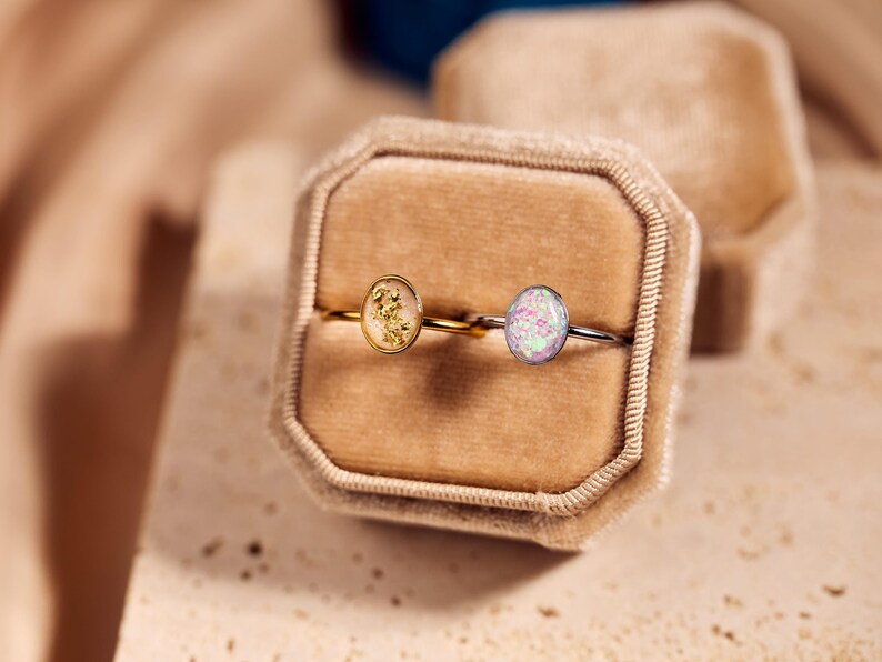 May include: Two oval-shaped rings displayed in a velvet ring box. One ring has a gold band with a gold-colored stone, and the other has a silver band with a light purple and pink stone. The box is beige.