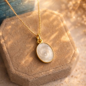 May include: A gold-colored necklace with an oval pendant. The pendant has a white, swirling design and a gold-colored border. The necklace is displayed on a beige velvet jewelry box.
