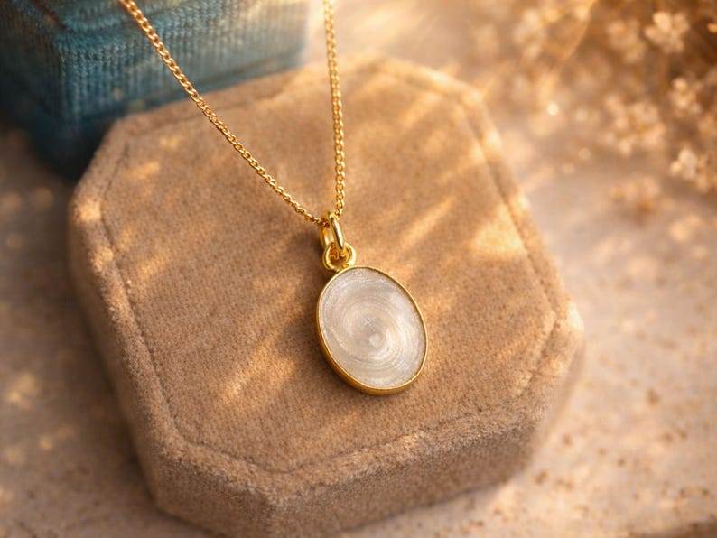 May include: A gold-colored necklace with an oval pendant. The pendant has a white, swirling design and a gold-colored border. The necklace is displayed on a beige velvet jewelry box.