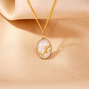 May include: A gold-colored necklace with an oval pendant. The pendant has a white background with gold flakes and a gold-colored border. The necklace chain is also gold-colored, and the background is a light tan fabric.