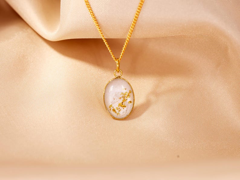 May include: A gold-colored necklace with an oval pendant. The pendant has a white background with gold flakes and a gold-colored border. The necklace chain is also gold-colored, and the background is a light tan fabric.