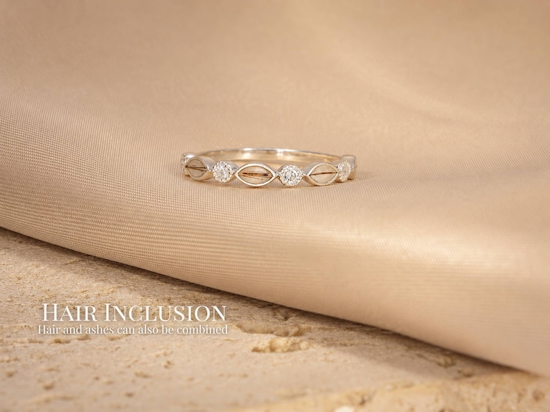 May include: A silver ring with a repeating pattern of oval shapes and small, round, clear stones. The ring is set against a beige fabric background. The text "HAIR INCLUSION" and "Hair and ashes can also be combined" is visible.