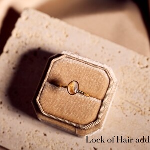 May include: A gold ring with an oval-shaped, amber-colored stone, presented in a velvet ring box. The ring is delicate, with a thin band. The text "Lock of Hair added" is visible at the bottom of the image.