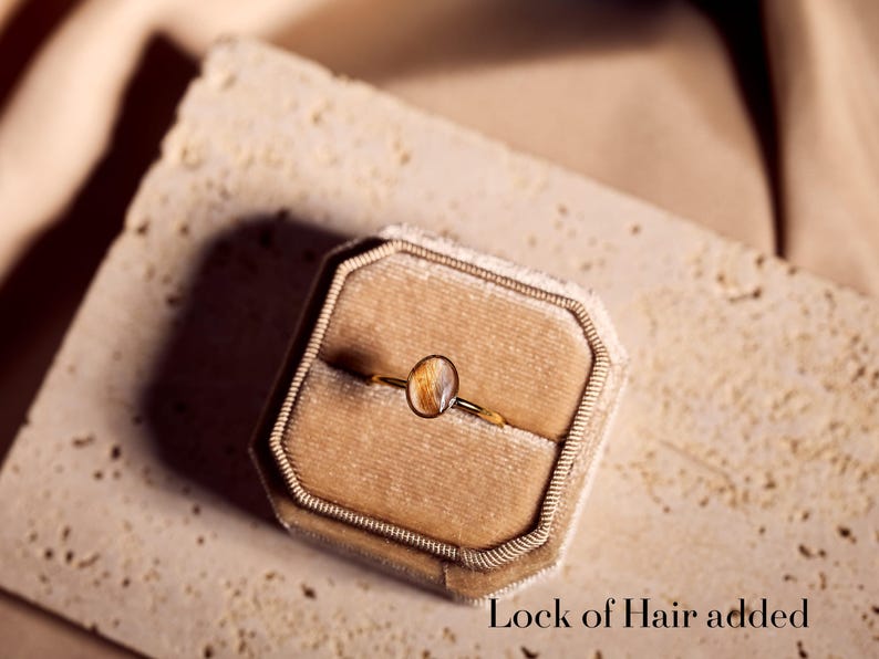 May include: A gold ring with an oval-shaped, amber-colored stone, presented in a velvet ring box. The ring is delicate, with a thin band. The text "Lock of Hair added" is visible at the bottom of the image.