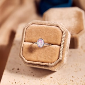May include: A silver ring with an oval-shaped, pink and white opal stone. The ring is displayed in a tan velvet ring box. The background features a soft, neutral color palette.