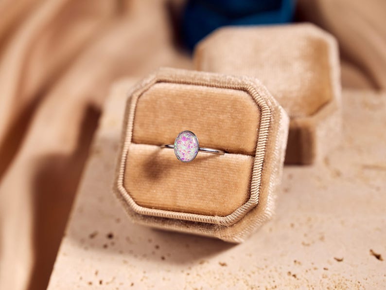 May include: A silver ring with an oval-shaped, pink and white opal stone. The ring is displayed in a tan velvet ring box. The background features a soft, neutral color palette.