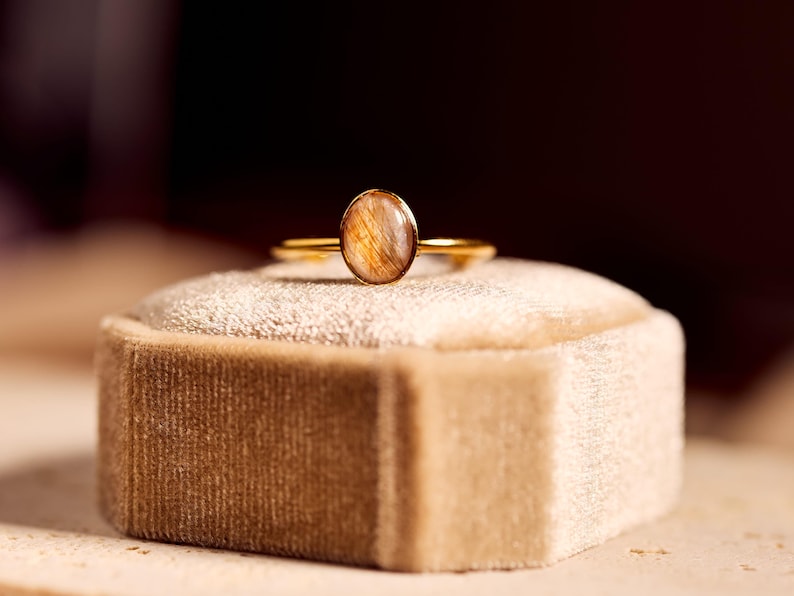 May include: A gold ring with an oval-shaped, light brown gemstone. The ring is set on a beige velvet ring box. The gemstone has a unique, natural pattern. The ring's band is thin and delicate, adding to its elegant appearance.