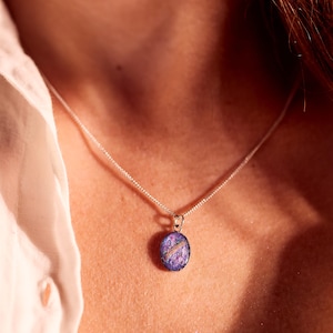May include: A silver necklace with a small, oval pendant. The pendant features a purple and blue design with a white accent. The necklace is worn against a white shirt.