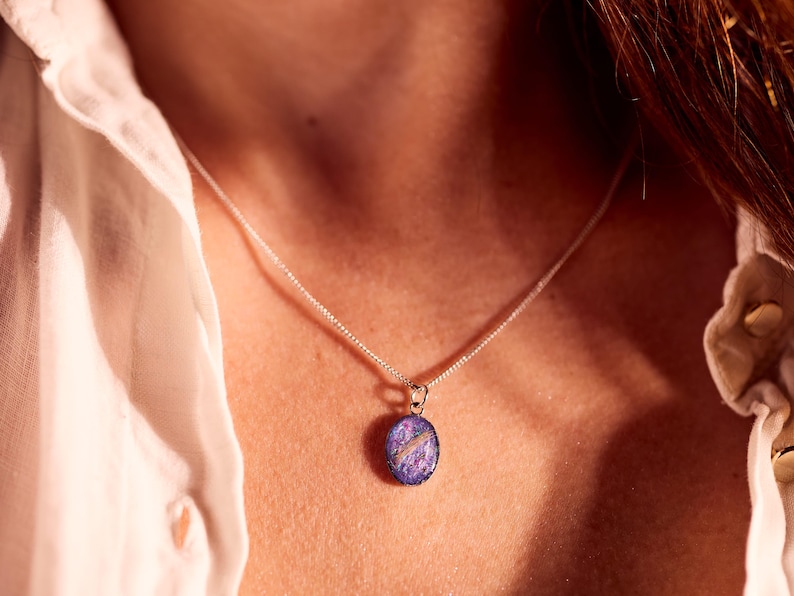 May include: A silver necklace with a small, oval pendant. The pendant features a purple and blue design with a white accent. The necklace is worn against a white shirt.