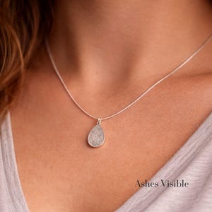 May include: A silver necklace with a teardrop-shaped pendant. The pendant has a textured, light gray surface. The necklace is worn against a light gray top. The text "Ashes Visible" is at the bottom right.