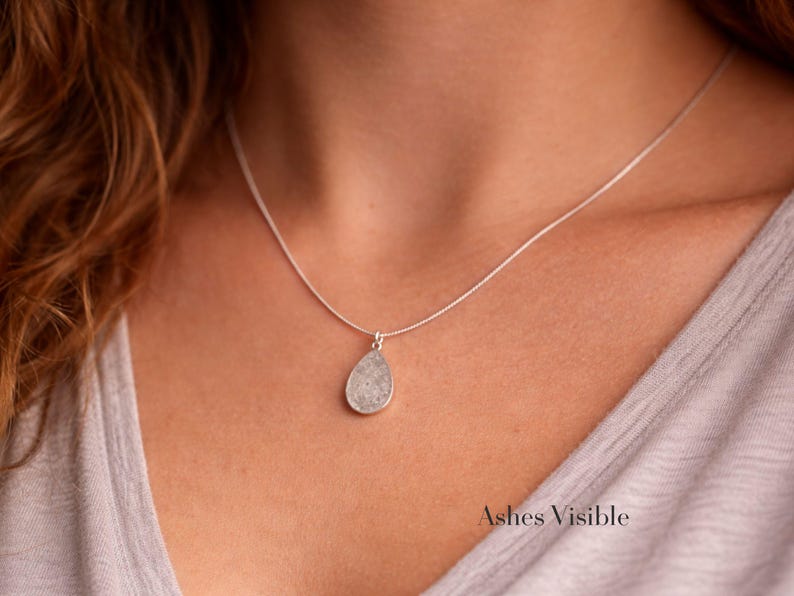 May include: A silver necklace with a teardrop-shaped pendant. The pendant has a textured, light gray surface. The necklace is worn against a light gray top. The text "Ashes Visible" is at the bottom right.