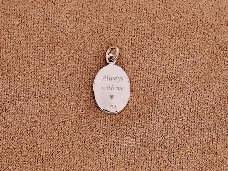 May include: Oval silver pendant with a loop at the top. The pendant is engraved with the words "Always with me" and a small heart. The pendant is on a brown textured surface.