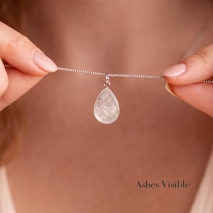 May include: A silver necklace with a teardrop-shaped pendant. The pendant is a clear, textured stone. The necklace is held by two hands. The text "Ashes Visible" is at the bottom of the image.