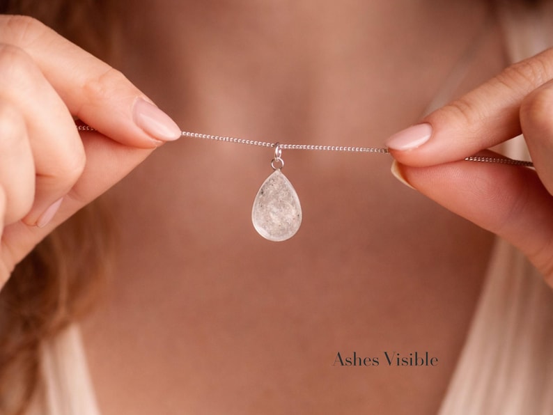 May include: A silver necklace with a teardrop-shaped pendant. The pendant is a clear, textured stone. The necklace is held by two hands. The text "Ashes Visible" is at the bottom of the image.