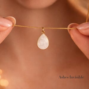 May include: A gold-colored necklace with a teardrop-shaped pendant. The pendant is white with a pearlescent sheen. The necklace is held by two hands. The text "Ashes Invisible" is at the bottom right.