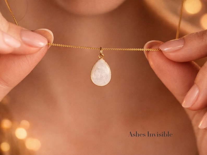 May include: A gold-colored necklace with a teardrop-shaped pendant. The pendant is white with a pearlescent sheen. The necklace is held by two hands. The text "Ashes Invisible" is at the bottom right.
