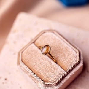 May include: A gold ring with an oval-shaped, light brown stone, set in a gold band. The ring is displayed in a beige velvet ring box. The background is a soft, pinkish-beige fabric.