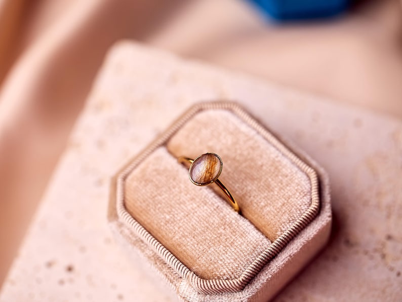 May include: A gold ring with an oval-shaped, light brown stone, set in a gold band. The ring is displayed in a beige velvet ring box. The background is a soft, pinkish-beige fabric.