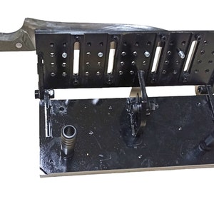 May include: A black metal knife sharpening jig with a large knife in place. The jig has multiple slots and holes, with a handle on the side. The knife has a wooden handle and a large blade.